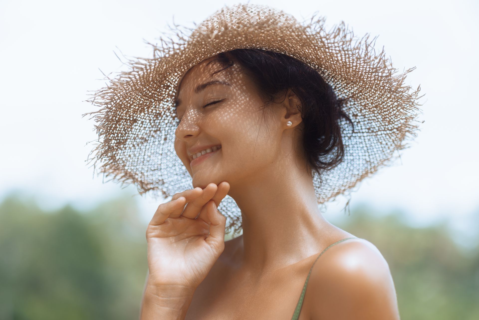 A woman wearing a straw hat is smiling and touching her face.