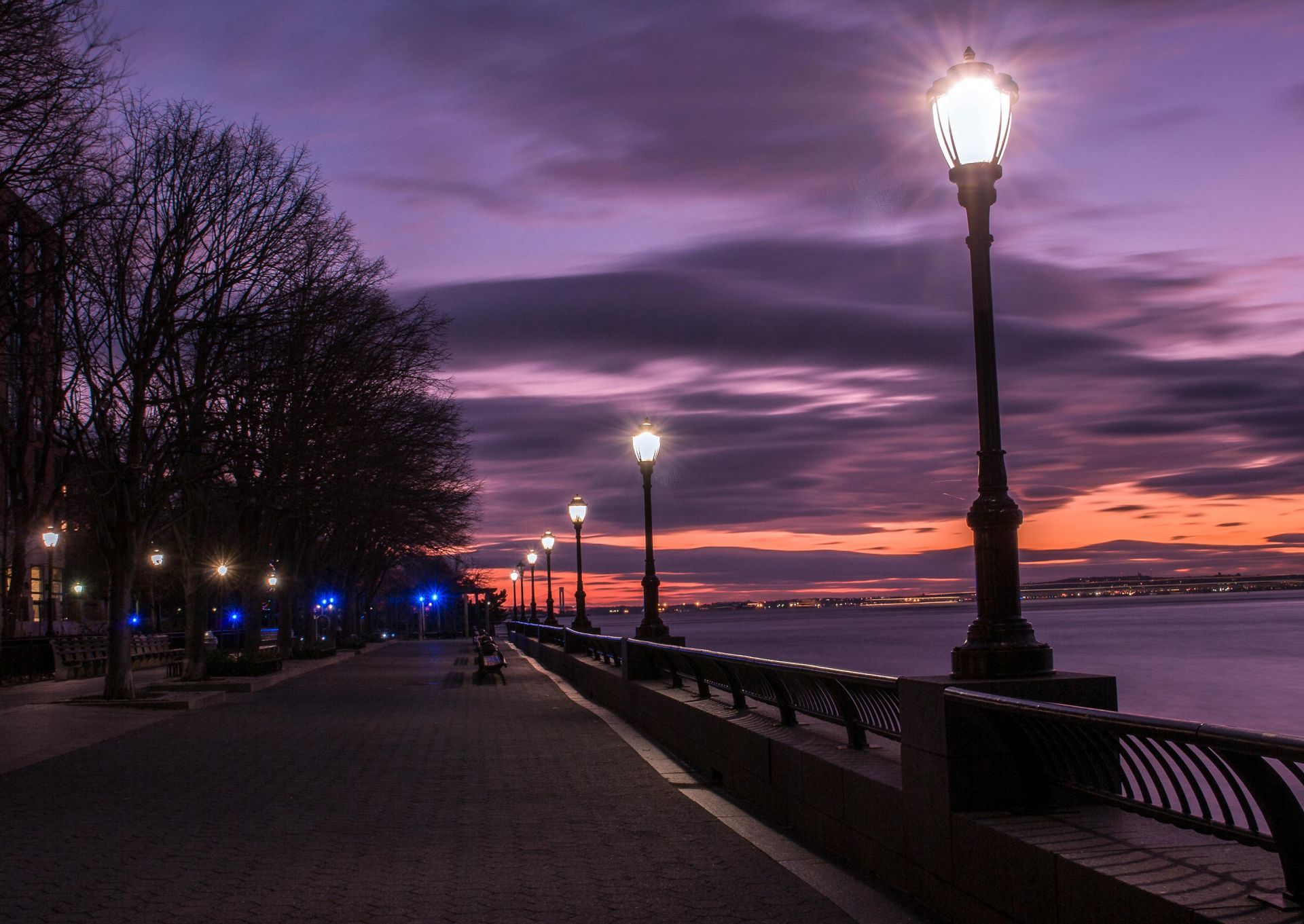 A row of street lights on a sidewalk next to a body of water at sunset.