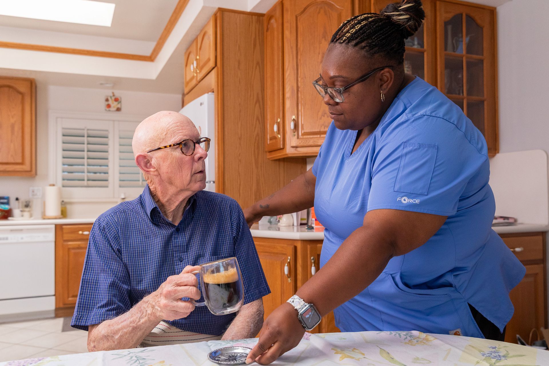 Caregiver assisting a person seated at a table, holding a mug, in a kitchen.