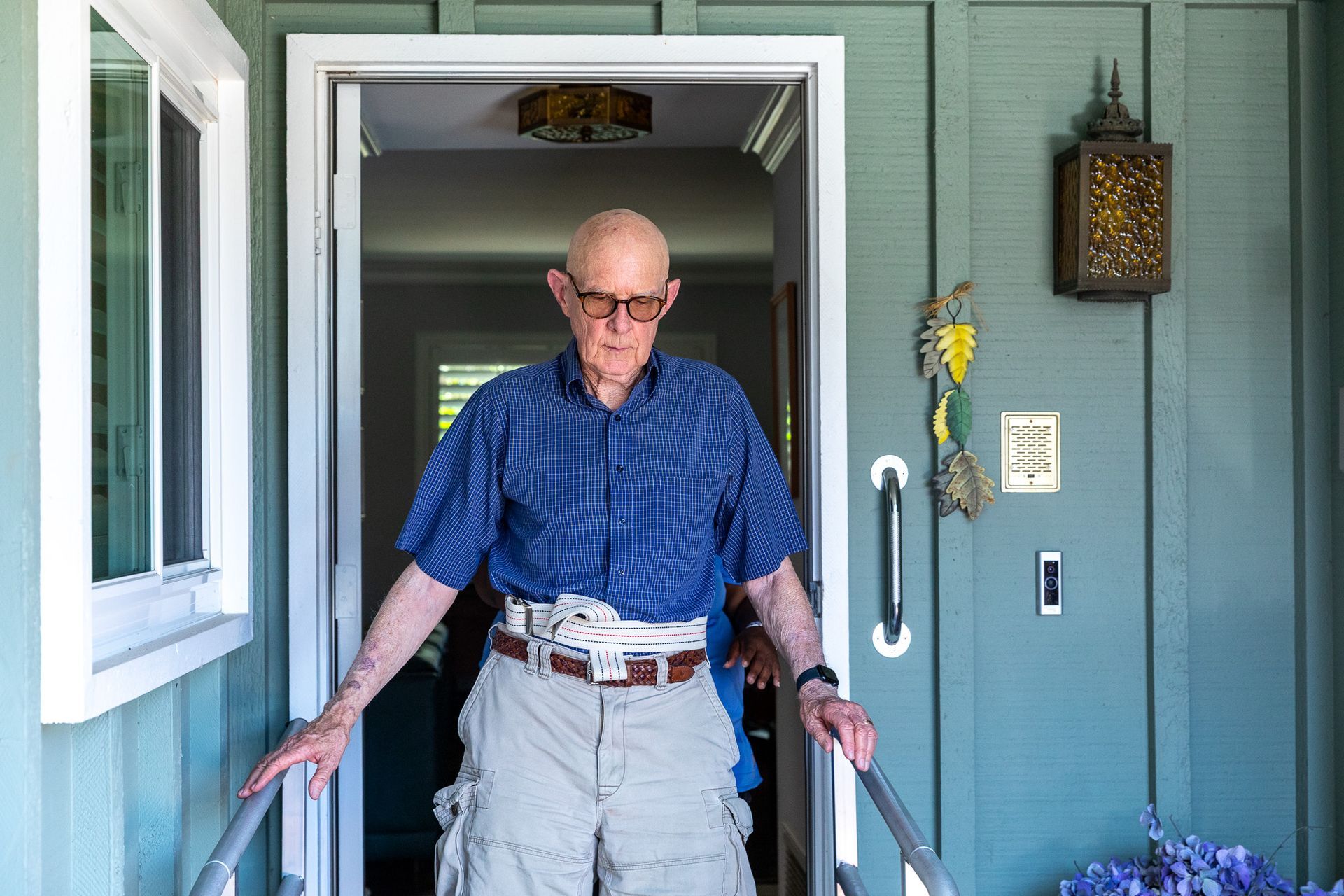 Man standing in a doorway with handrails, wearing a blue shirt and khaki shorts on a porch