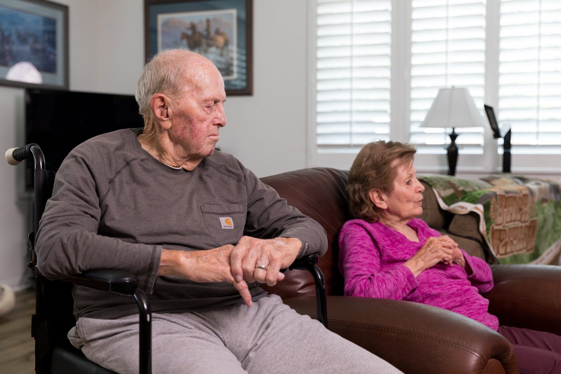 Older person in wheelchair, looking left, next to another person sitting on a sofa. Indoor setting.