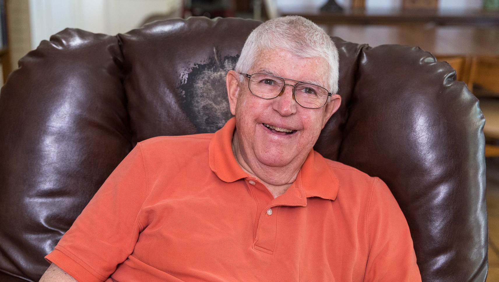 An elderly man is sitting in a chair wearing glasses and an orange shirt.
