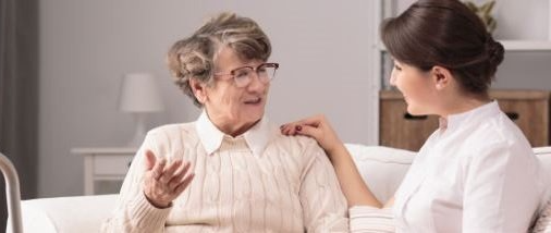 An elderly woman is sitting on a couch talking to a nurse.
