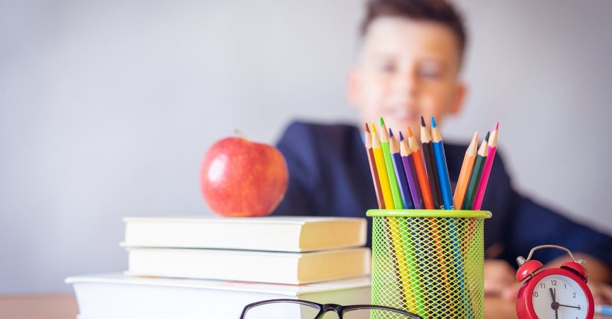 A boy is sitting at a desk with books , pencils , glasses and an apple.