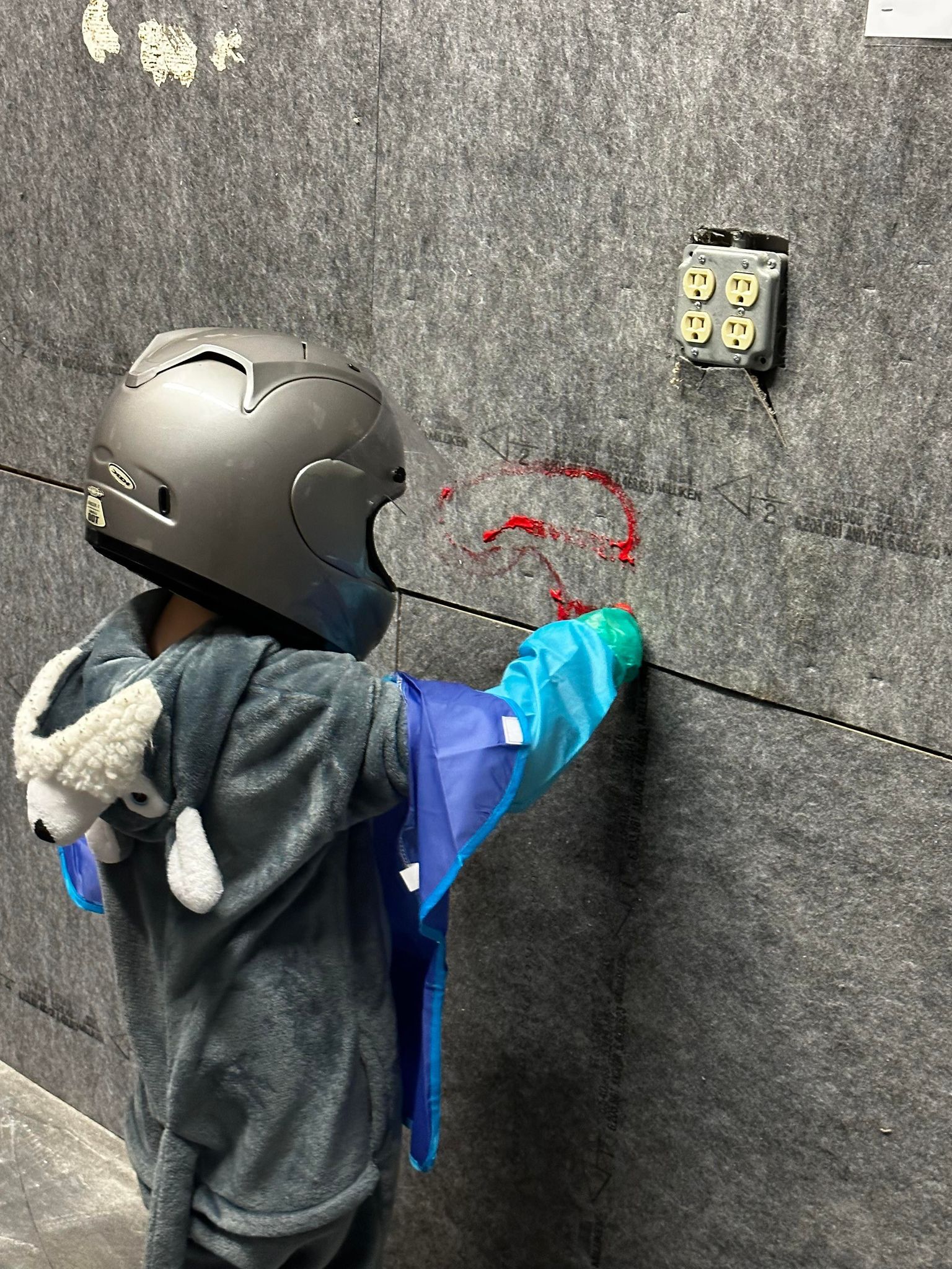 A young boy wearing a helmet and a cape is standing next to a wall.