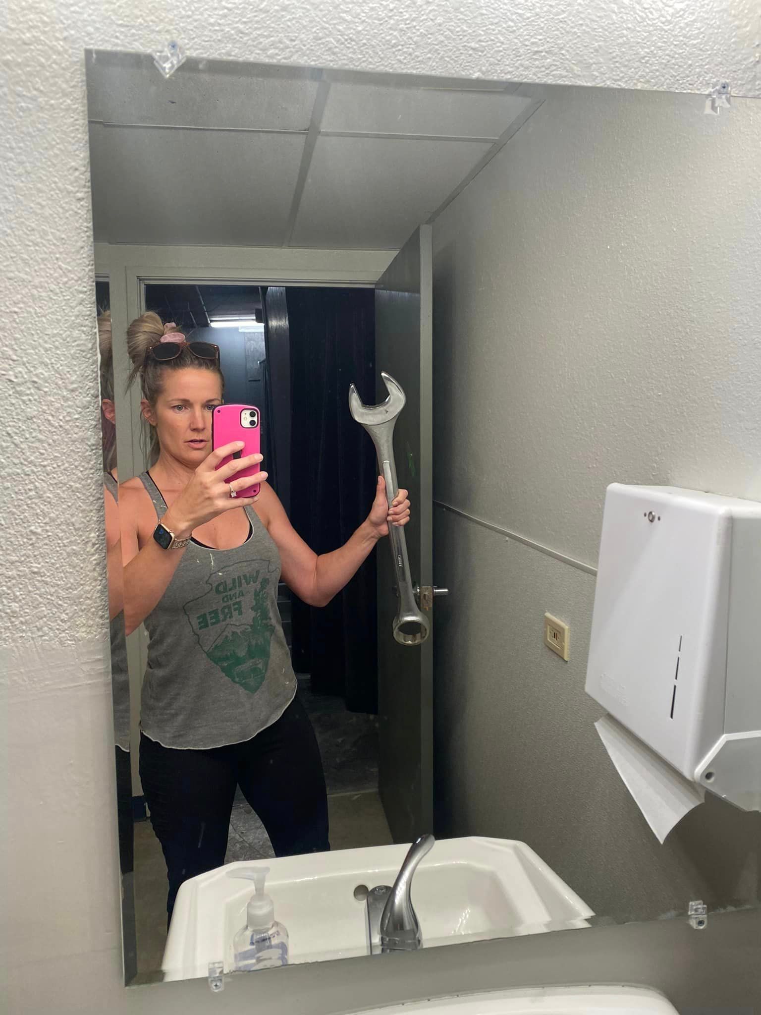 A woman is taking a selfie in a bathroom mirror while holding a wrench.