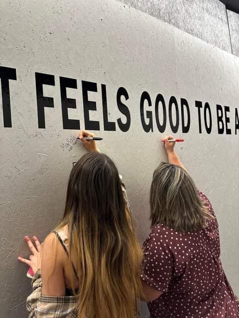 Two women are writing on a wall that says it feels good to be a