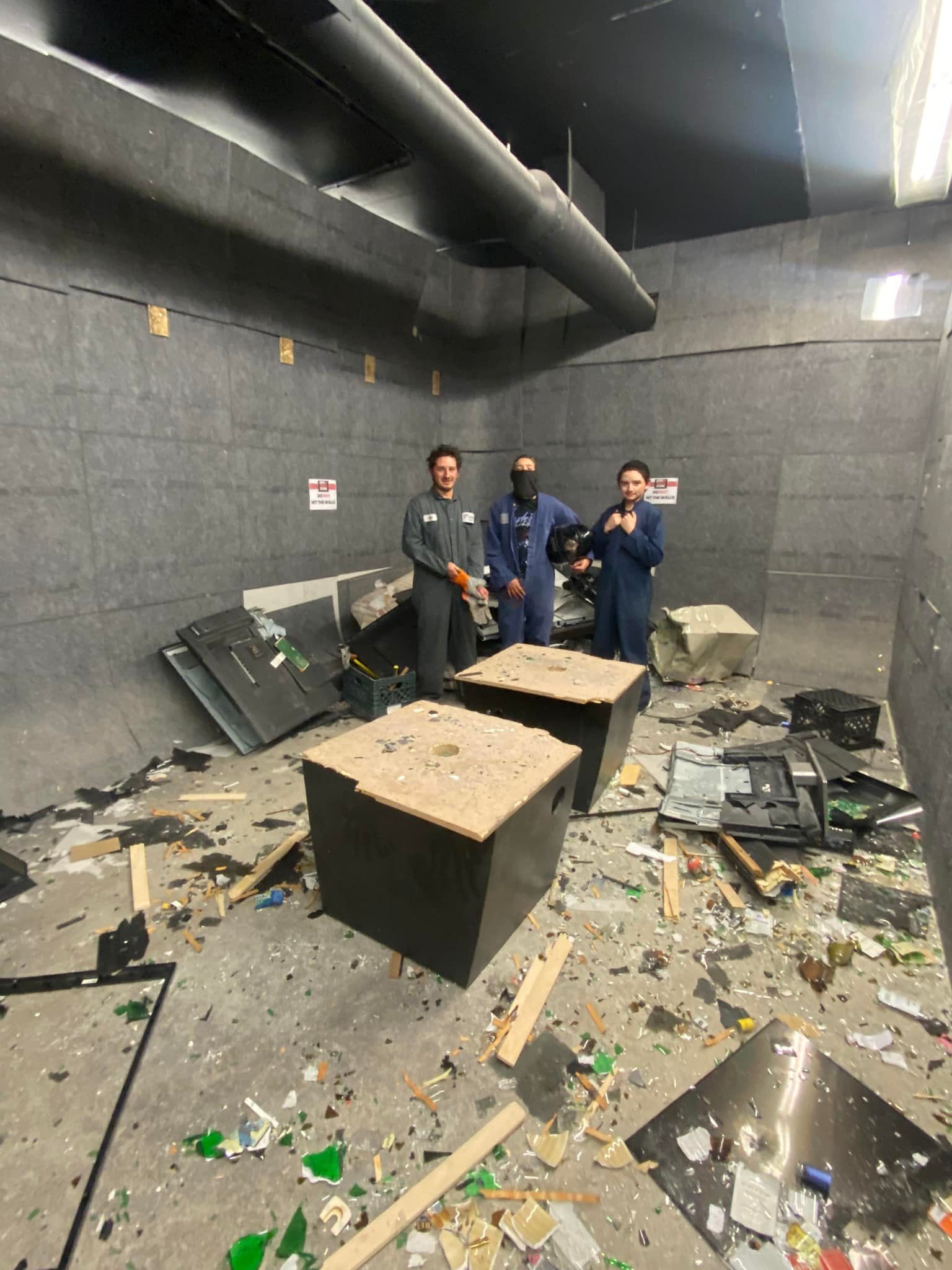 Three men are standing in a room with a lot of broken glass on the floor.