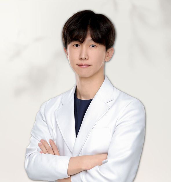 A person wearing a white lab coat and round-rimmed glasses against a light gray background.