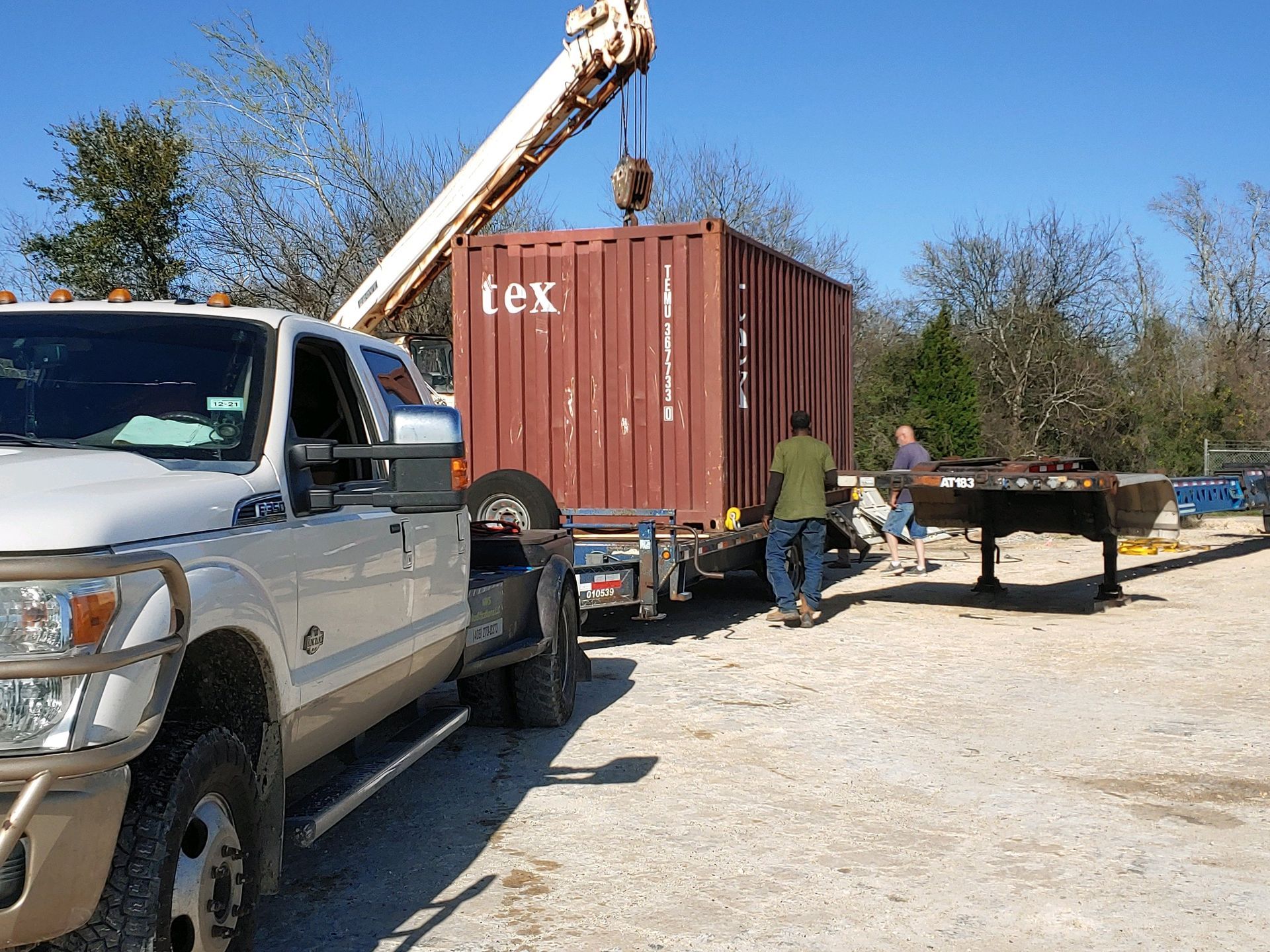 A crane lifting a red shipping container off a flatbed trailer, with two men assisting, in an outdoor setting.