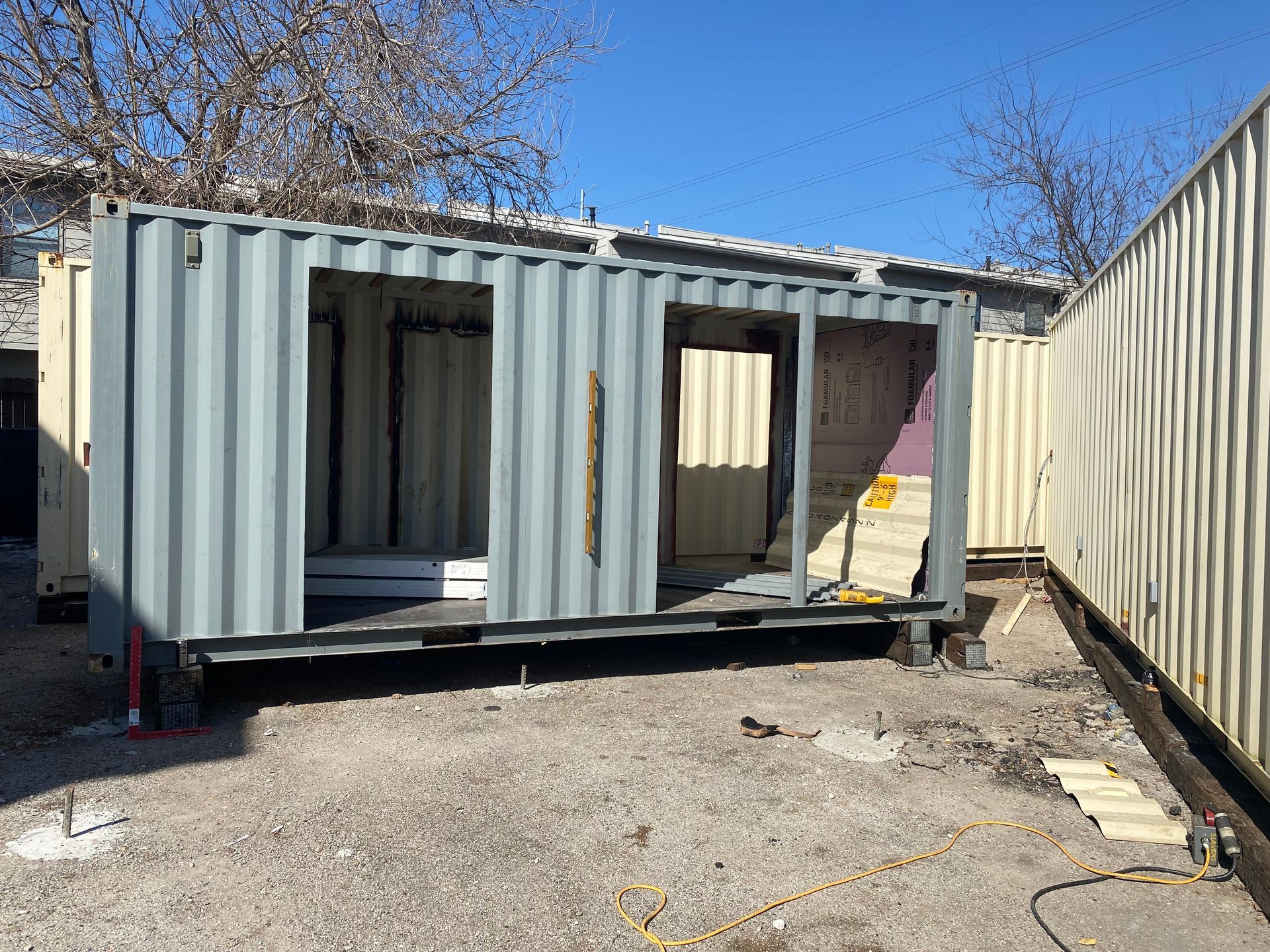 Gray shipping container with door/window frames cut out, in an outdoor setting.
