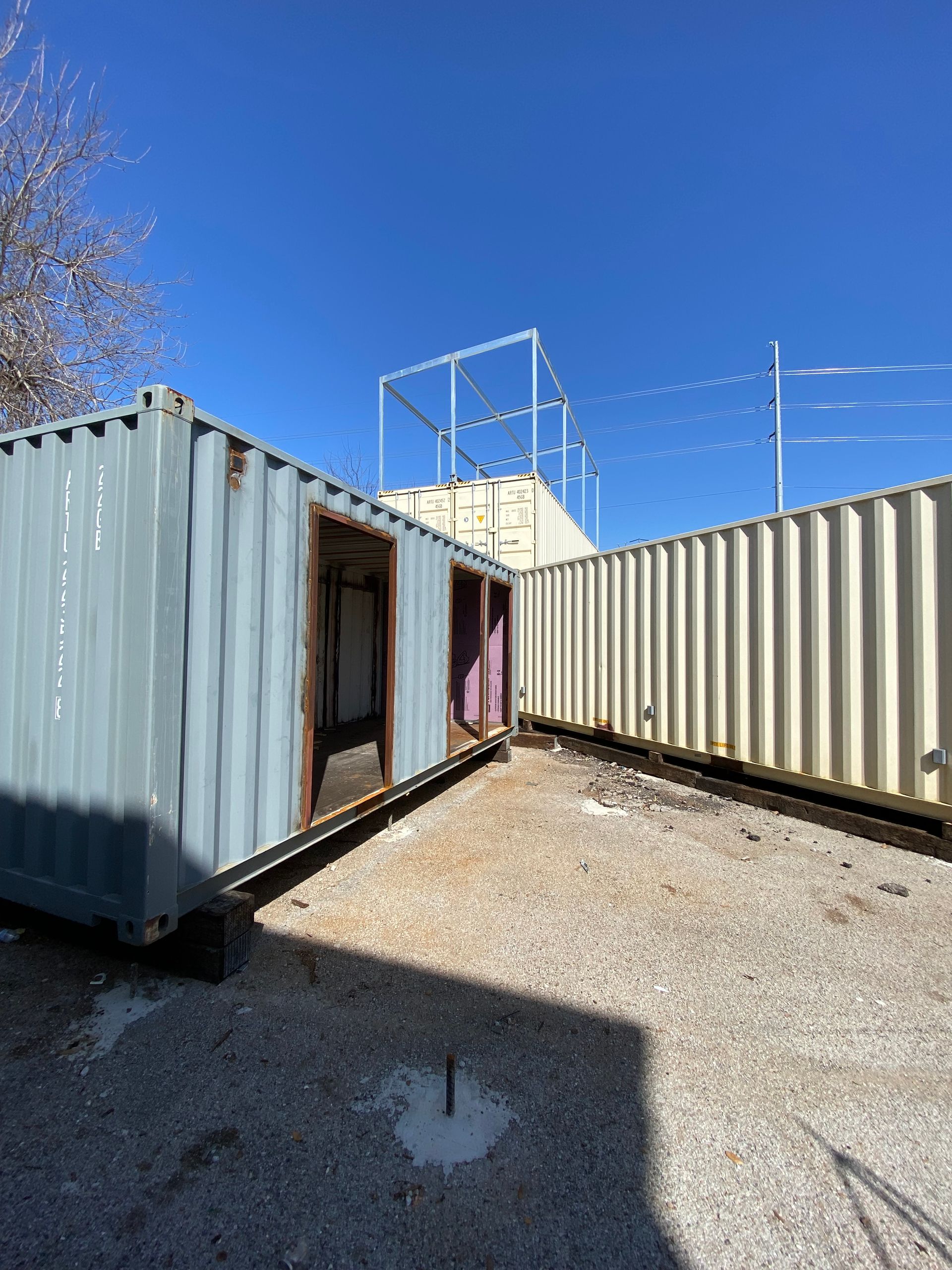 Two shipping containers, one blue-gray with open doors, another cream, sit on gravel under a blue sky.