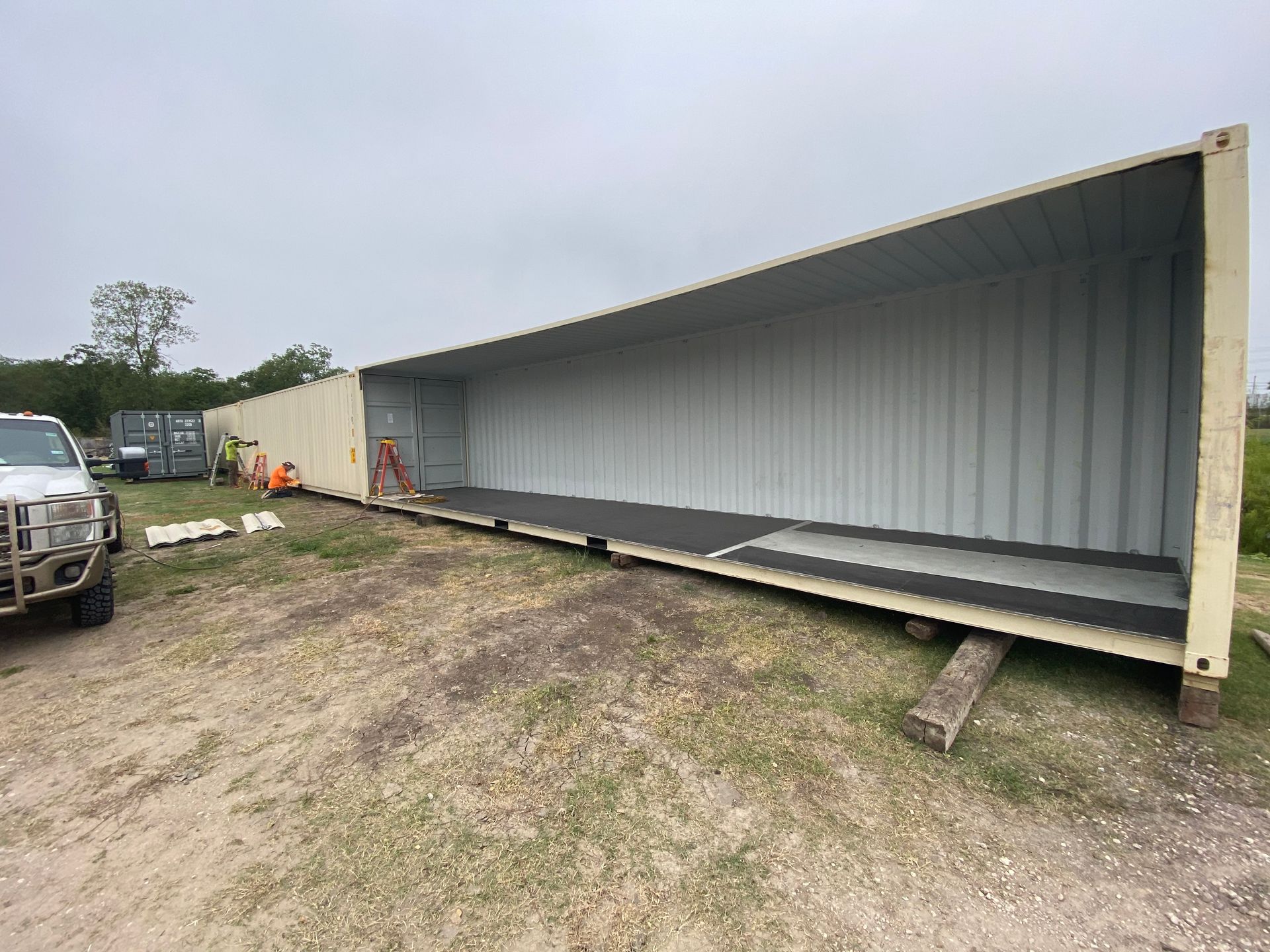 Empty, long shipping container with open top and workers nearby, outdoors on dirt.