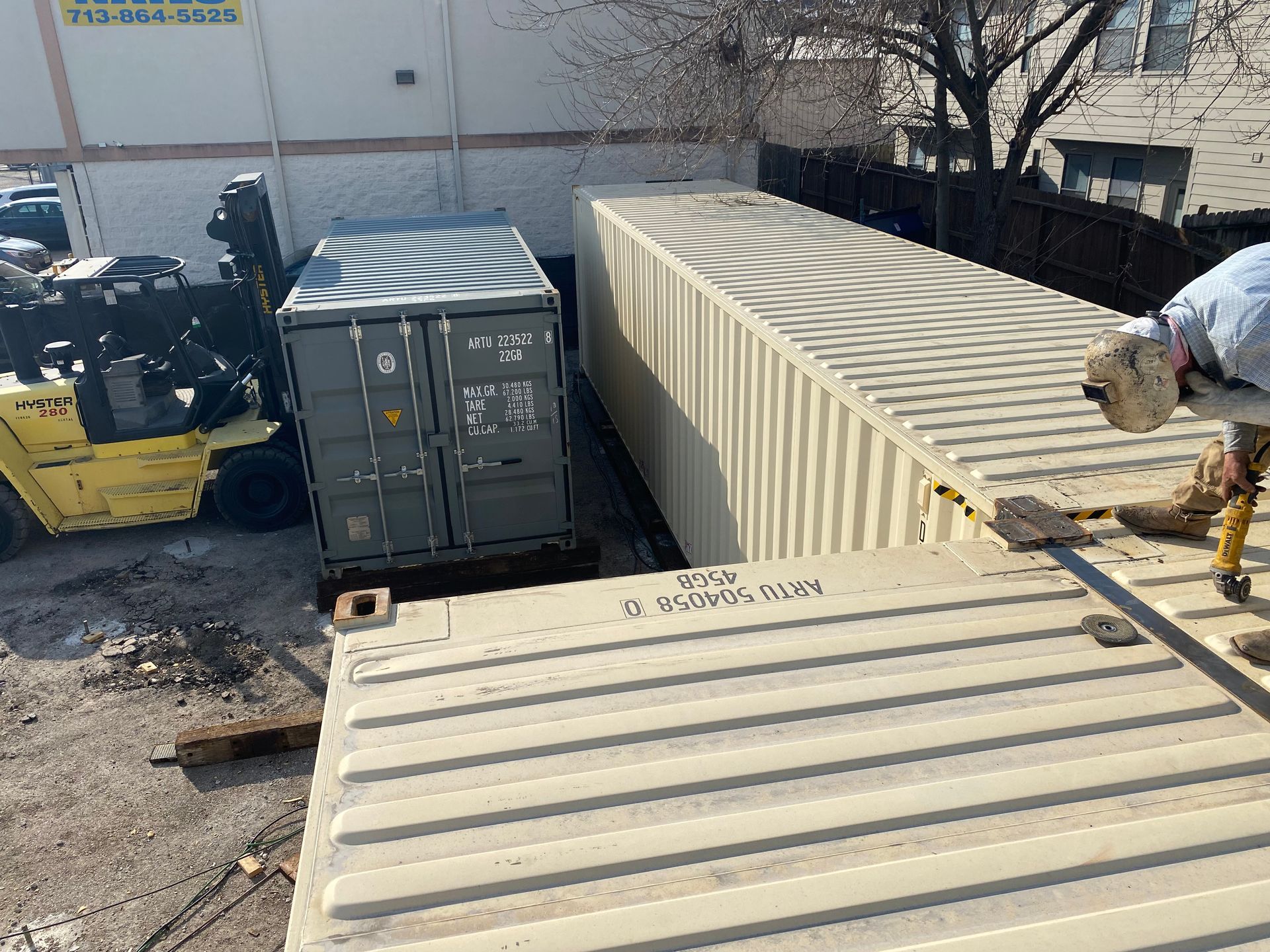 Forklift loading shipping containers; worker near one. Outdoors, sunny.