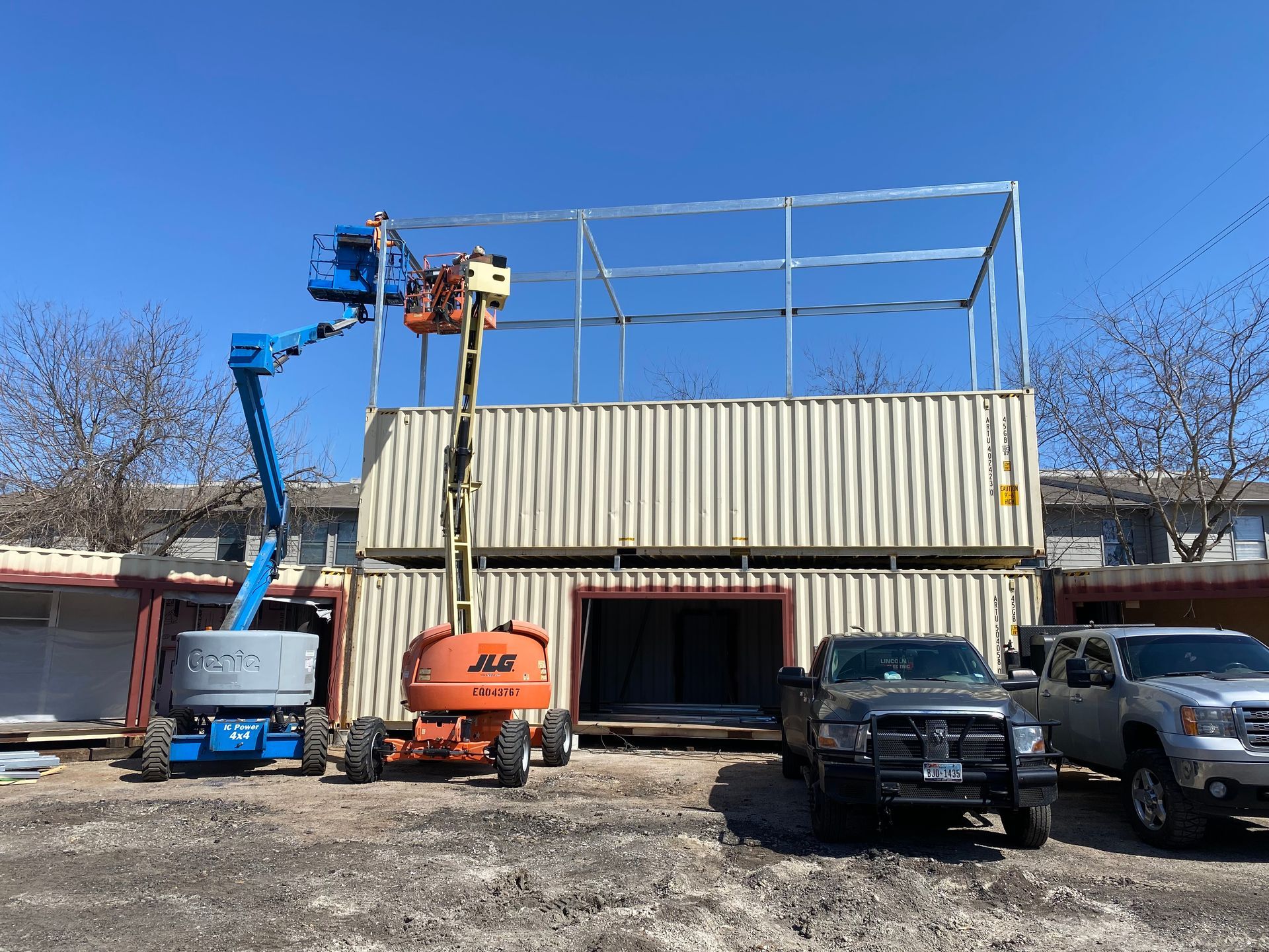 Two stacked shipping containers with a metal frame being built on top, workers on lifts.