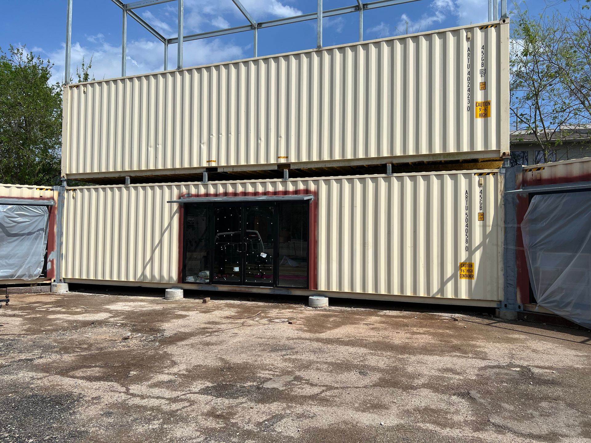 Two beige shipping containers stacked, one with glass door, outdoors.