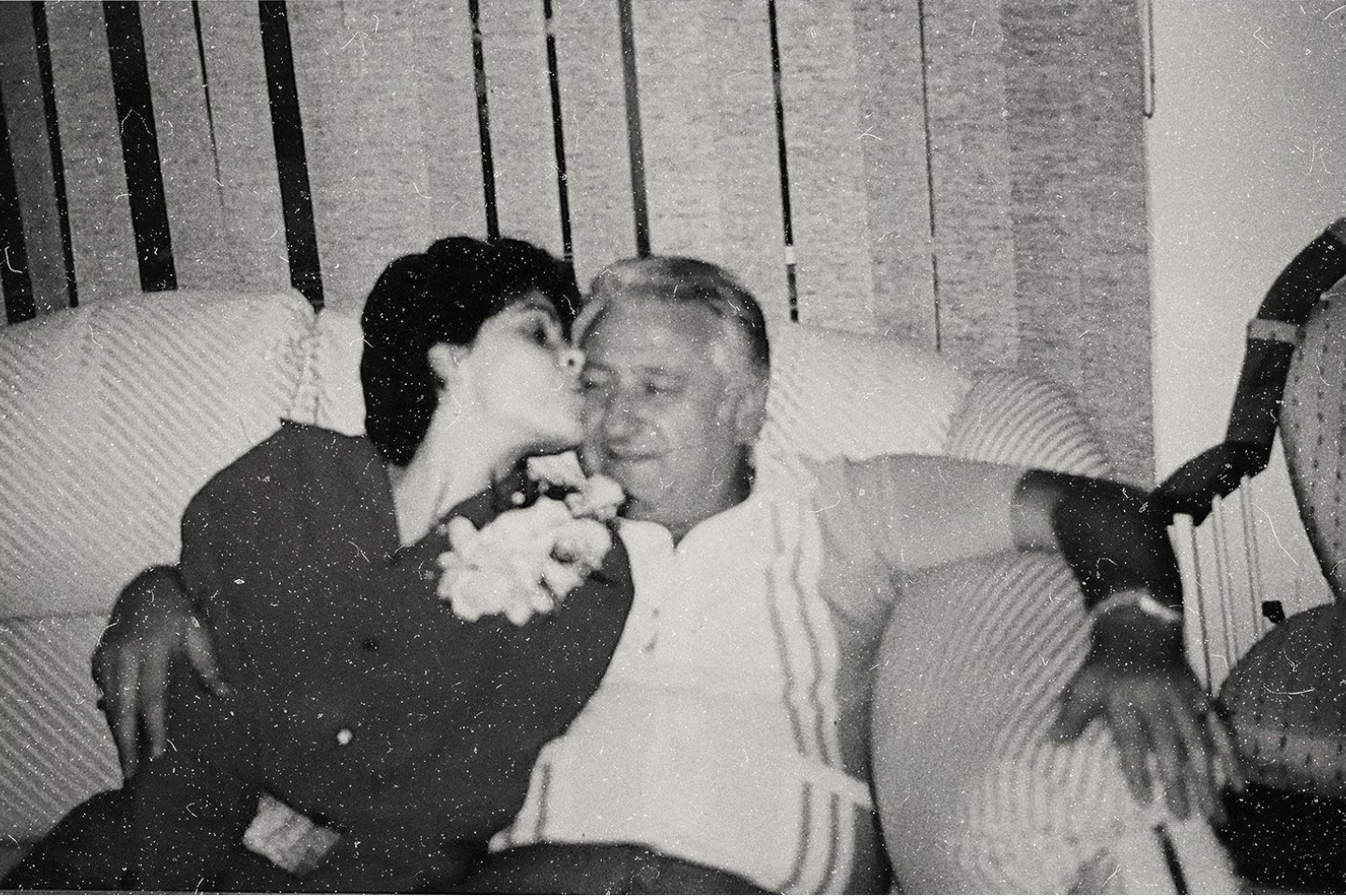 Woman kisses a man on the cheek; they sit on a couch. The man wears a polo, the woman holds flowers.