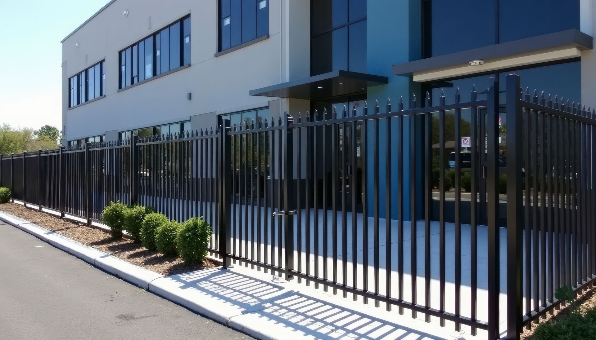 Black metal security fence surrounding a modern building with large windows; bushes along the fence. Black metal security fence surrounding a modern building with large windows; bushes along the fence.