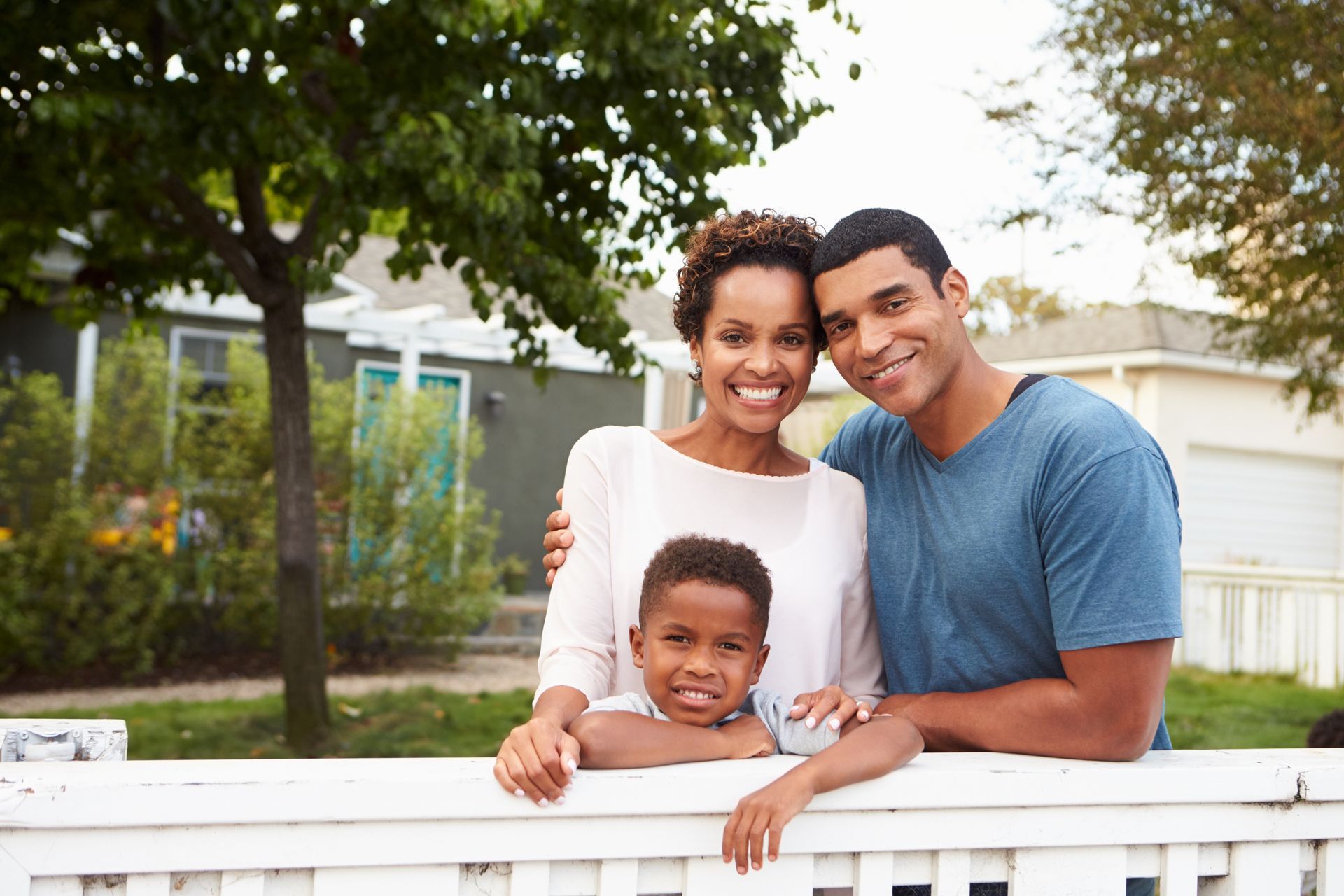 Family smiling in front of their home, leaning on a white fence. Family smiling in front of their home, leaning on a white fence.