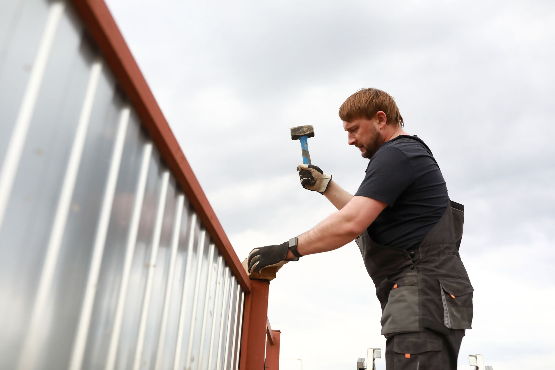 A person in work clothes hammering on a red metal structure, against a cloudy sky. A person in work clothes hammering on a red metal structure, against a cloudy sky.