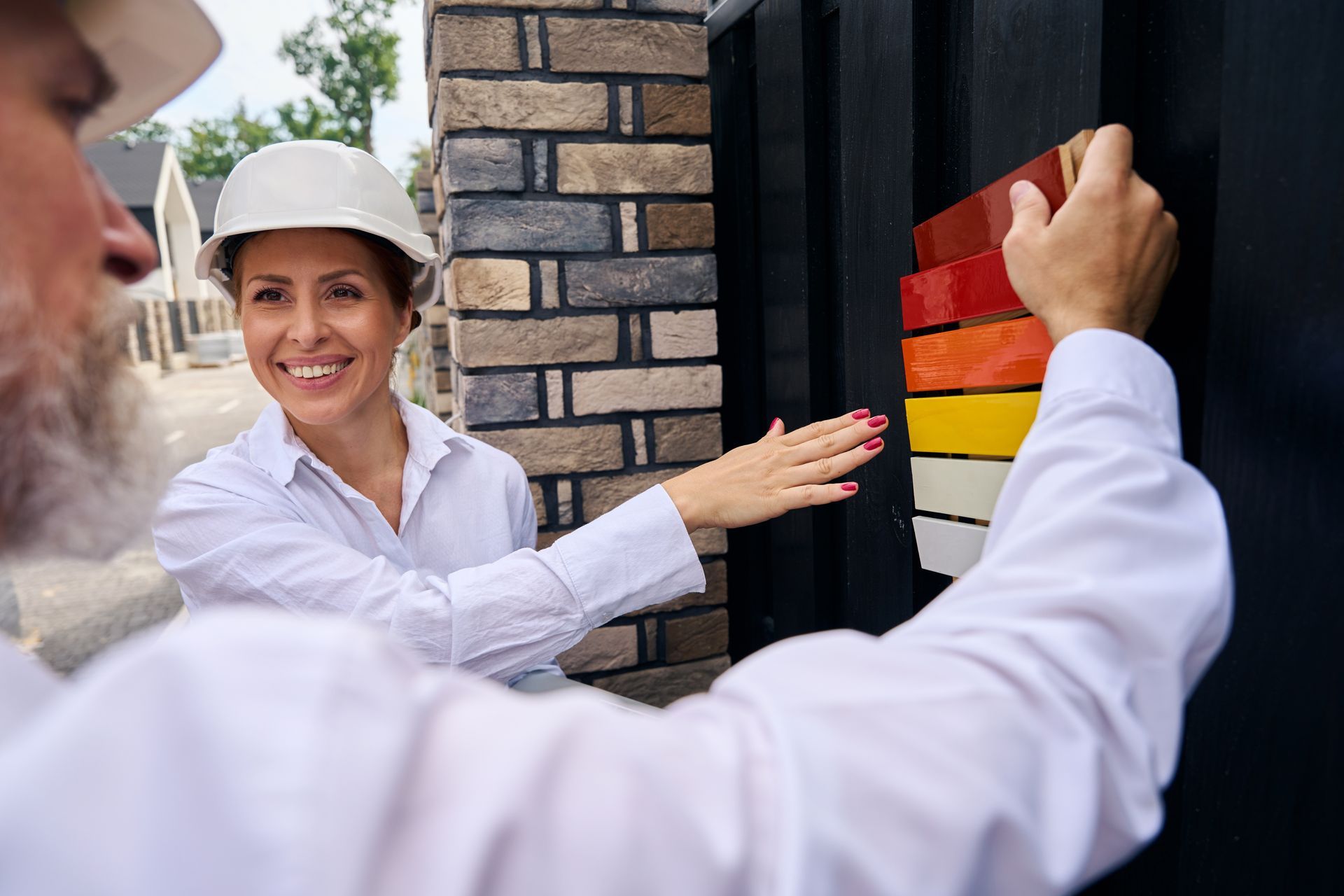 Two people in white shirts and hard hats reviewing paint swatches on a black structure next to a brick wall. Two people in white shirts and hard hats reviewing paint swatches on a black structure next to a brick wall.