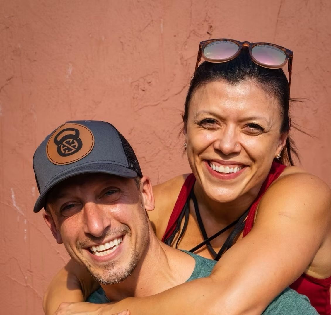Man wearing cap and woman smiling, embracing, against a coral wall.