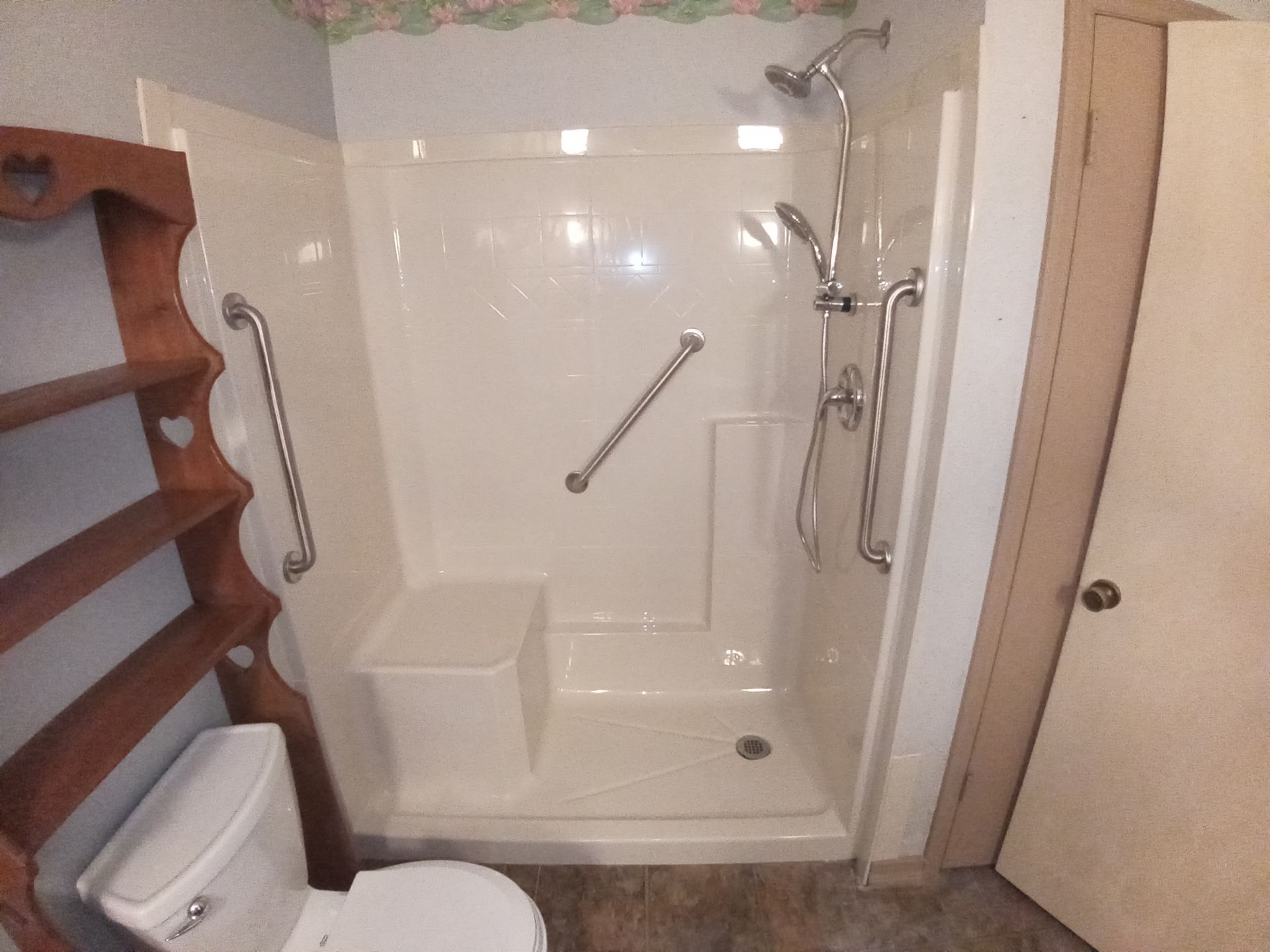A walk-in shower with grab bars and a built-in seat, situated next to a toilet and wooden shelving unit.