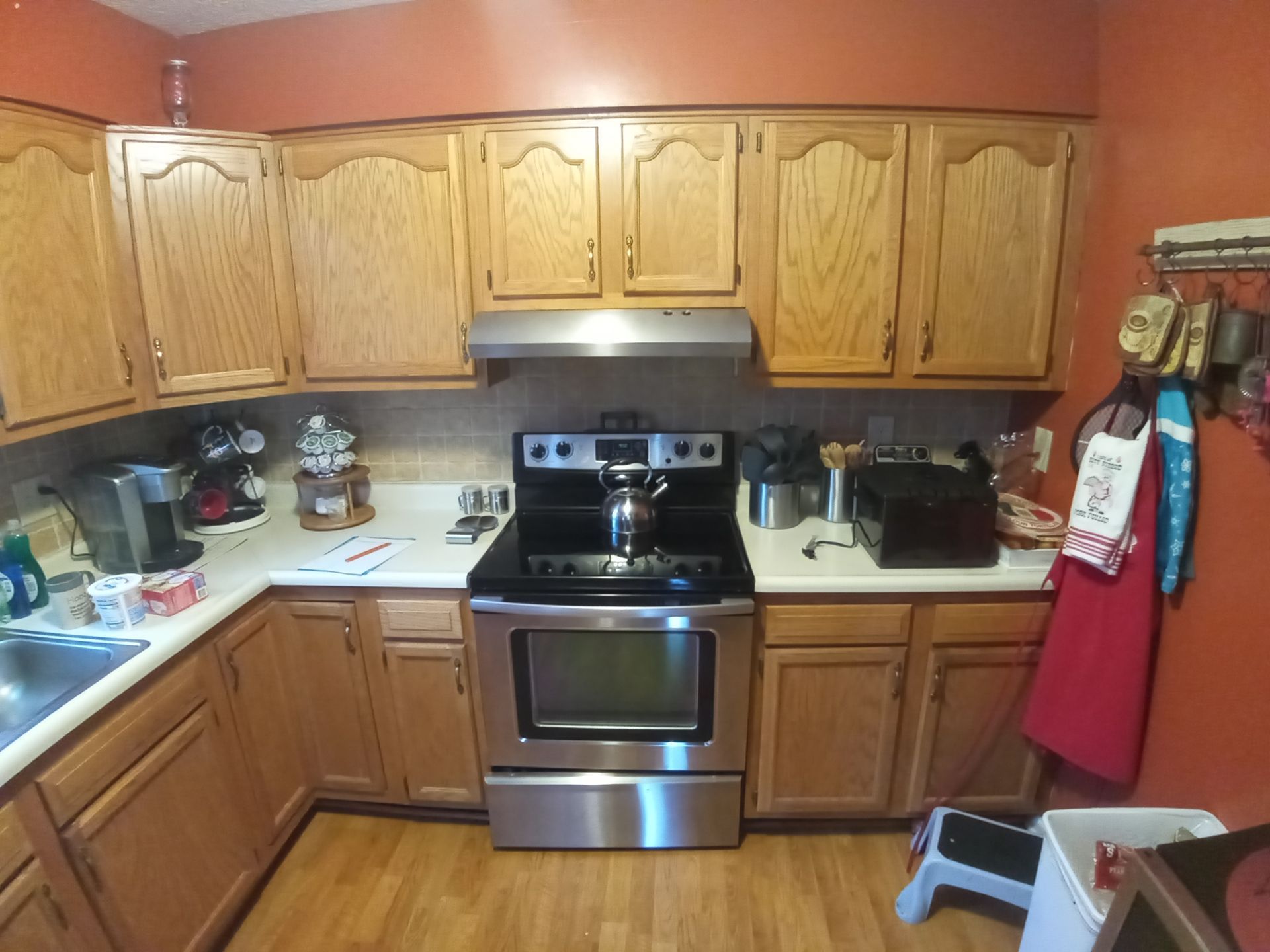A kitchen featuring light wood cabinets, an orange wall, a stainless steel stove with a kettle, and white countertops.