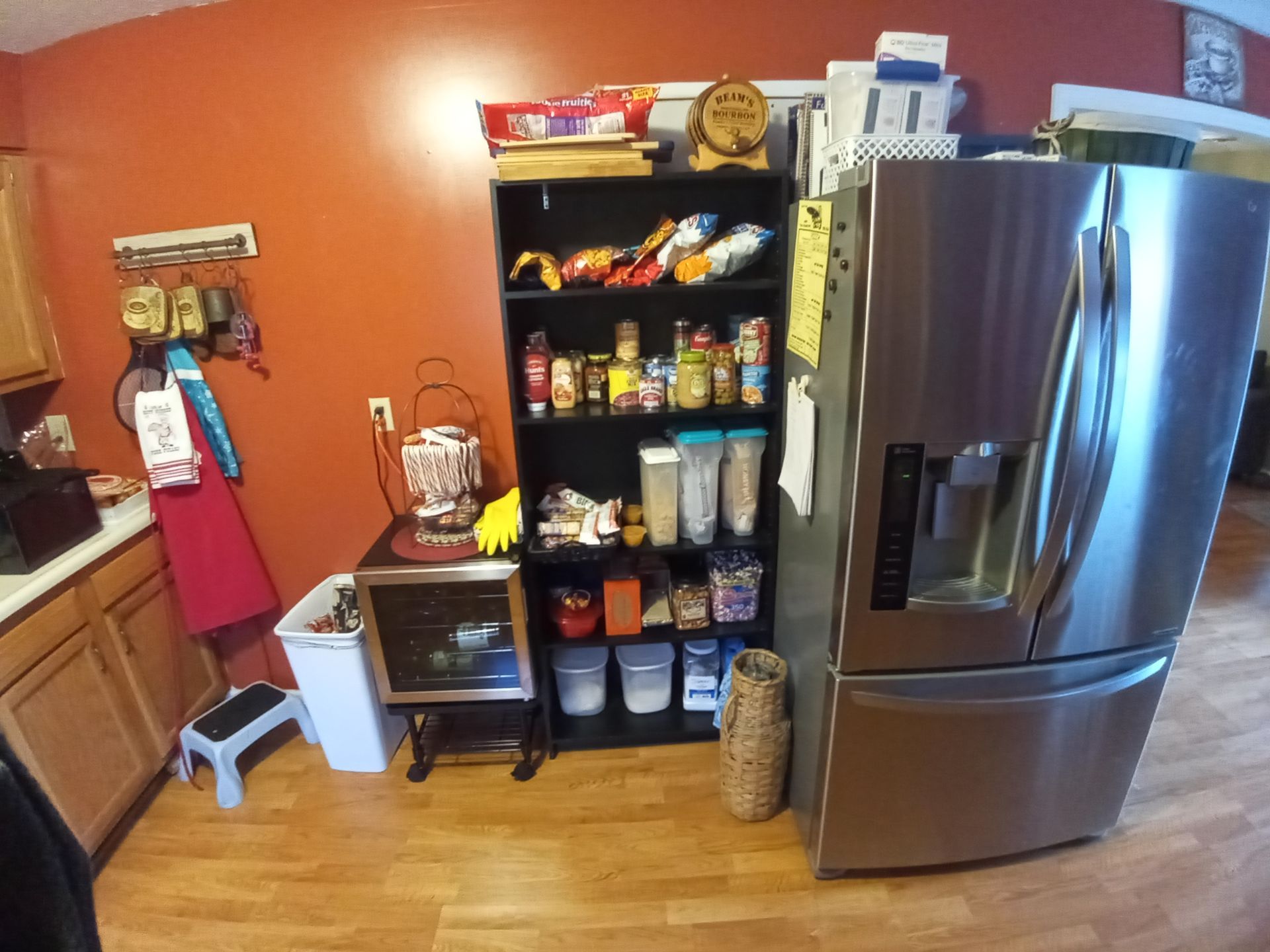 A tall, black pantry shelf filled with food stands next to a stainless steel refrigerator in a room with orange walls.