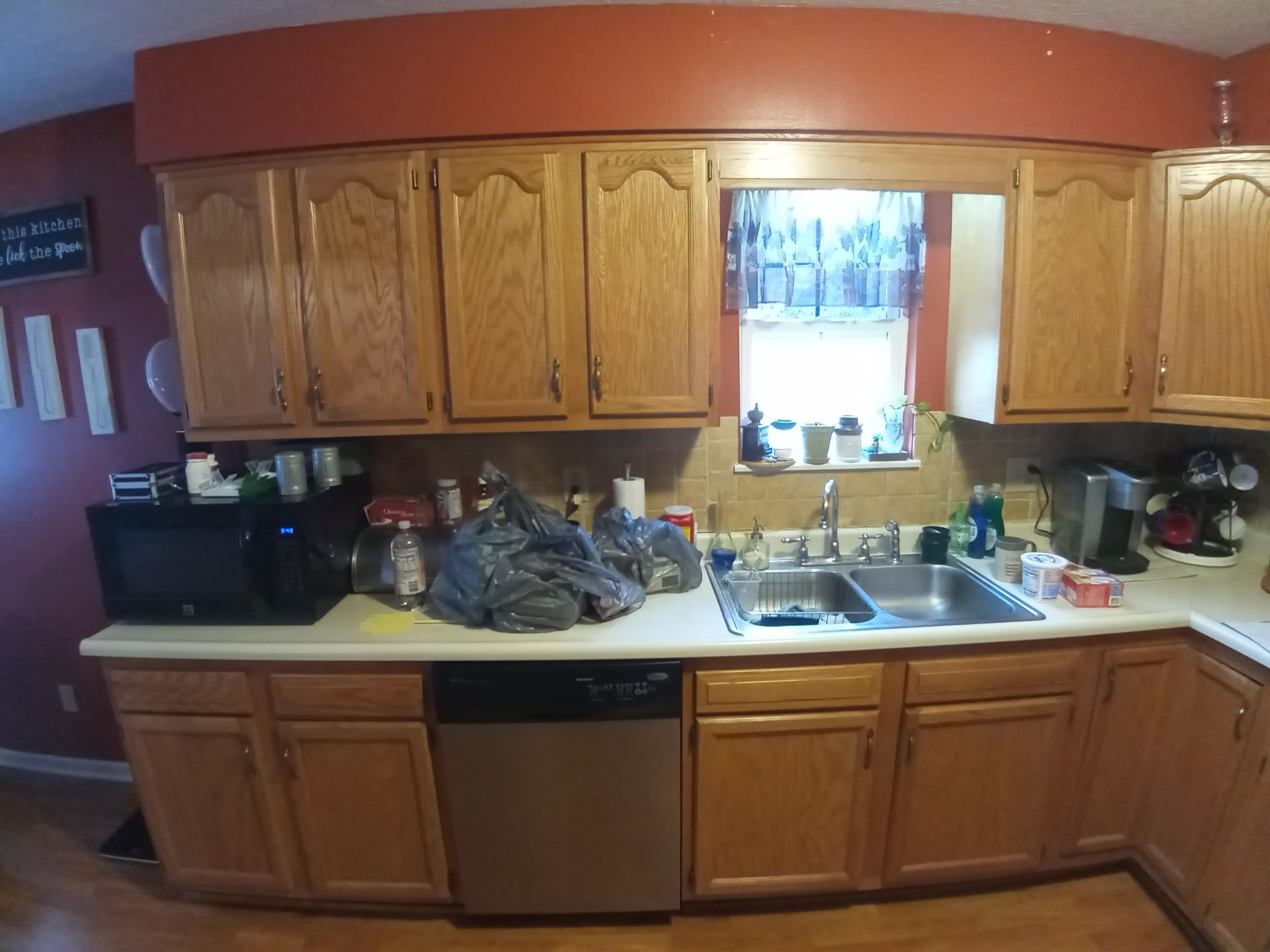 Kitchen sink and counters with oak cabinets, dark red walls, a microwave, dishwasher, and clutter on the countertops.
