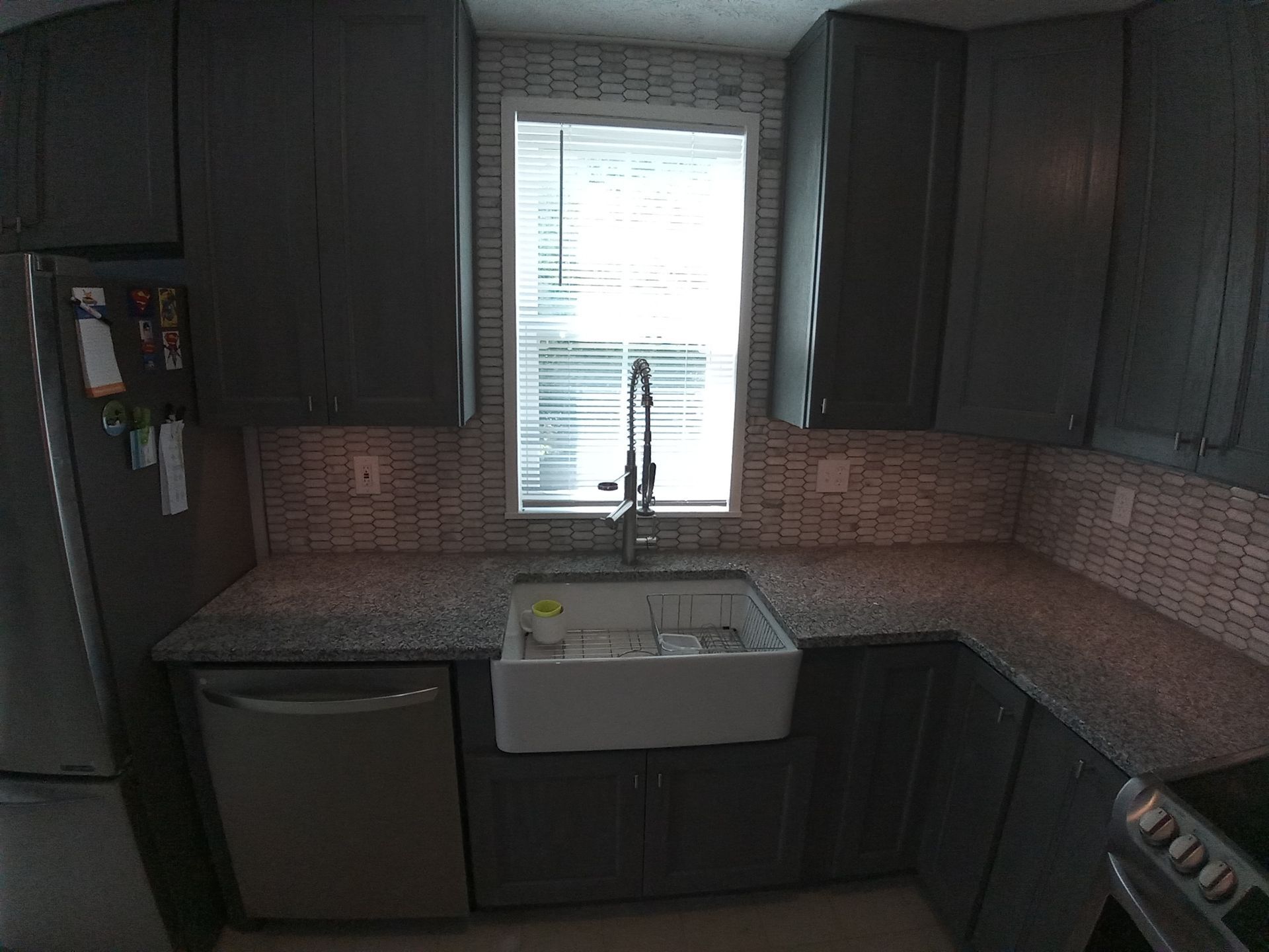 A dark grey kitchen with granite countertops, a white farmhouse sink, and a window with blinds above the sink.