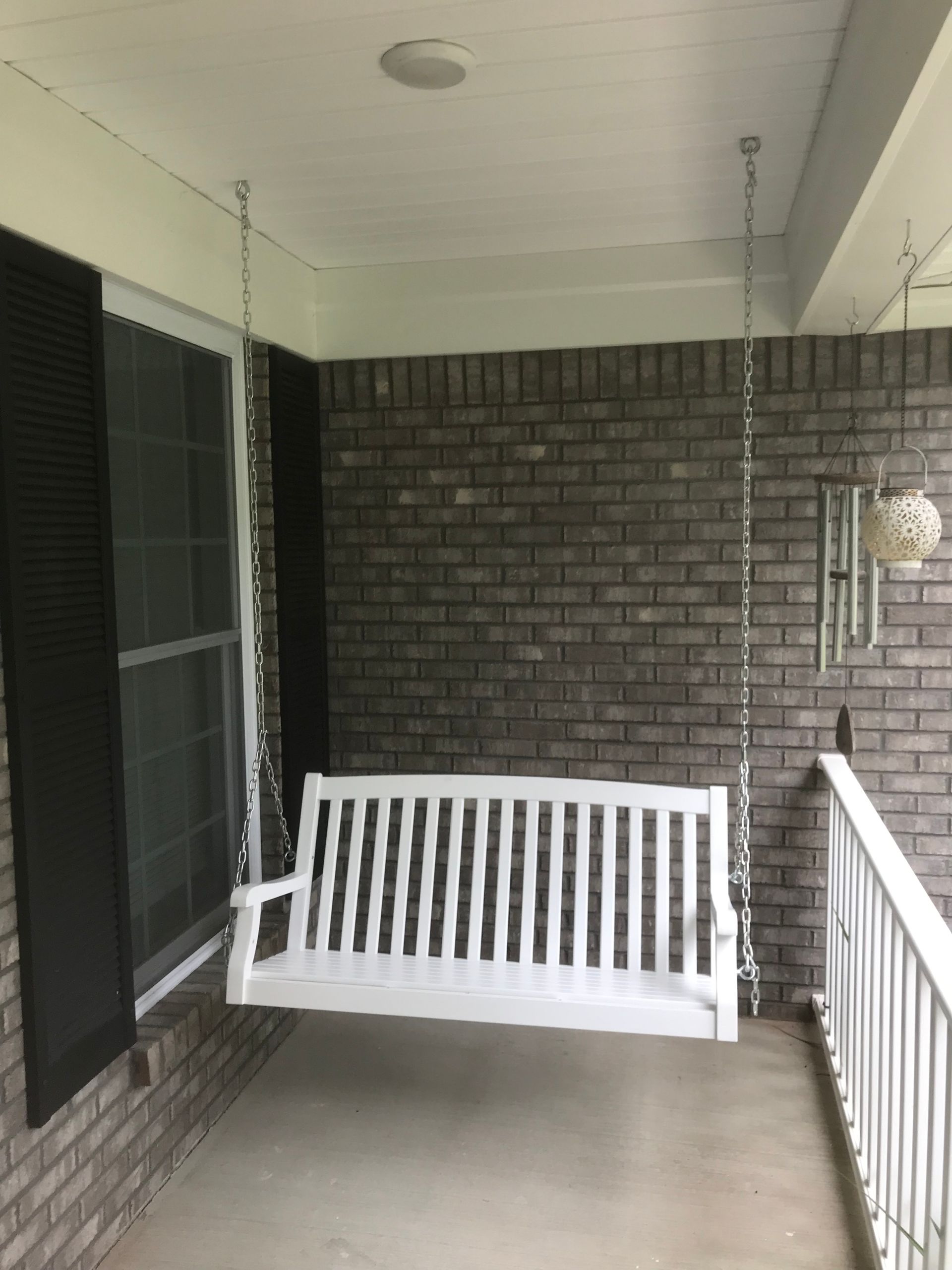 A white wooden porch swing hanging by metal chains from the ceiling of a covered porch with dark brick walls.