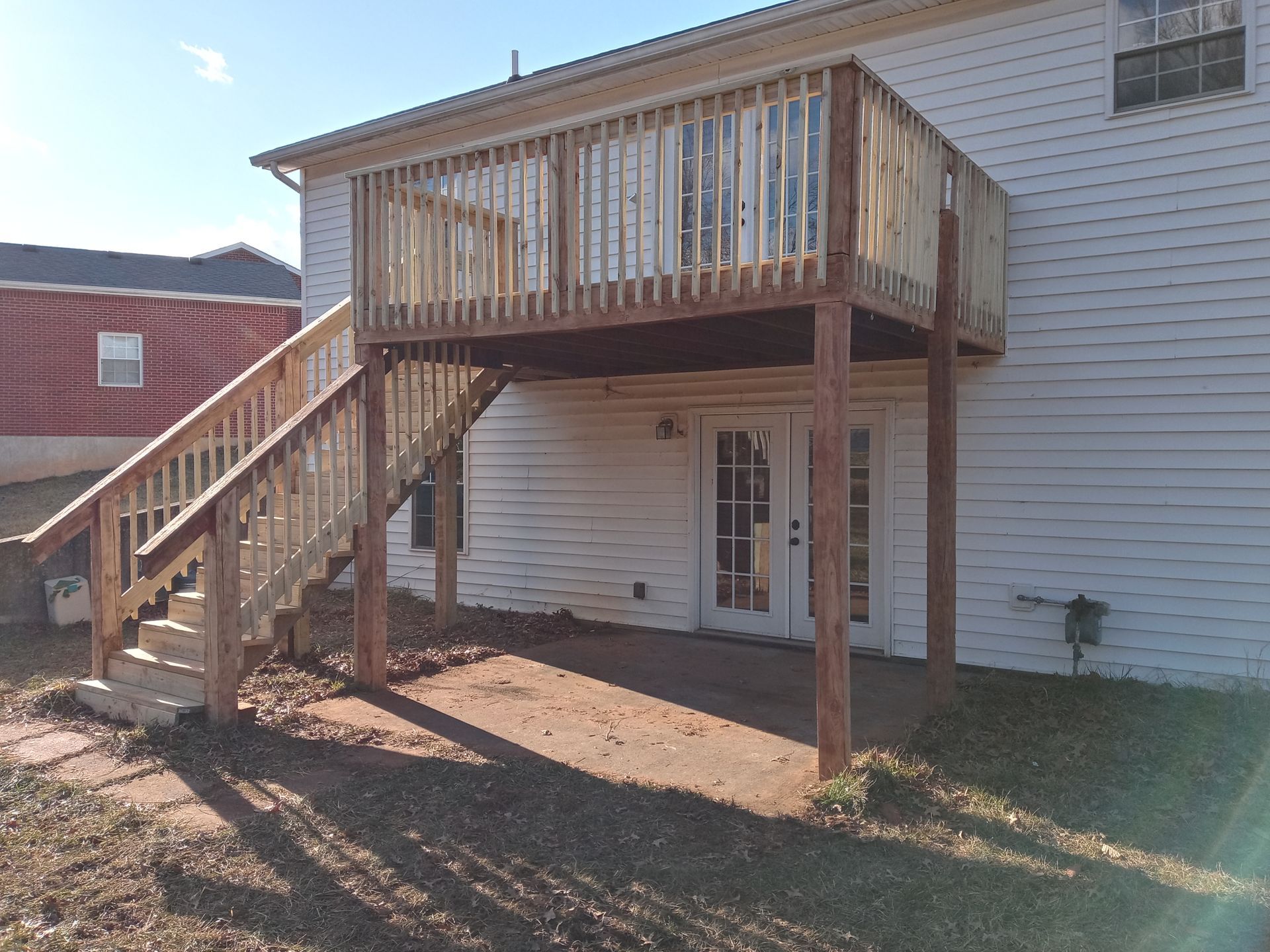 A wooden deck with a staircase leading down to a concrete patio behind a white-sided house.