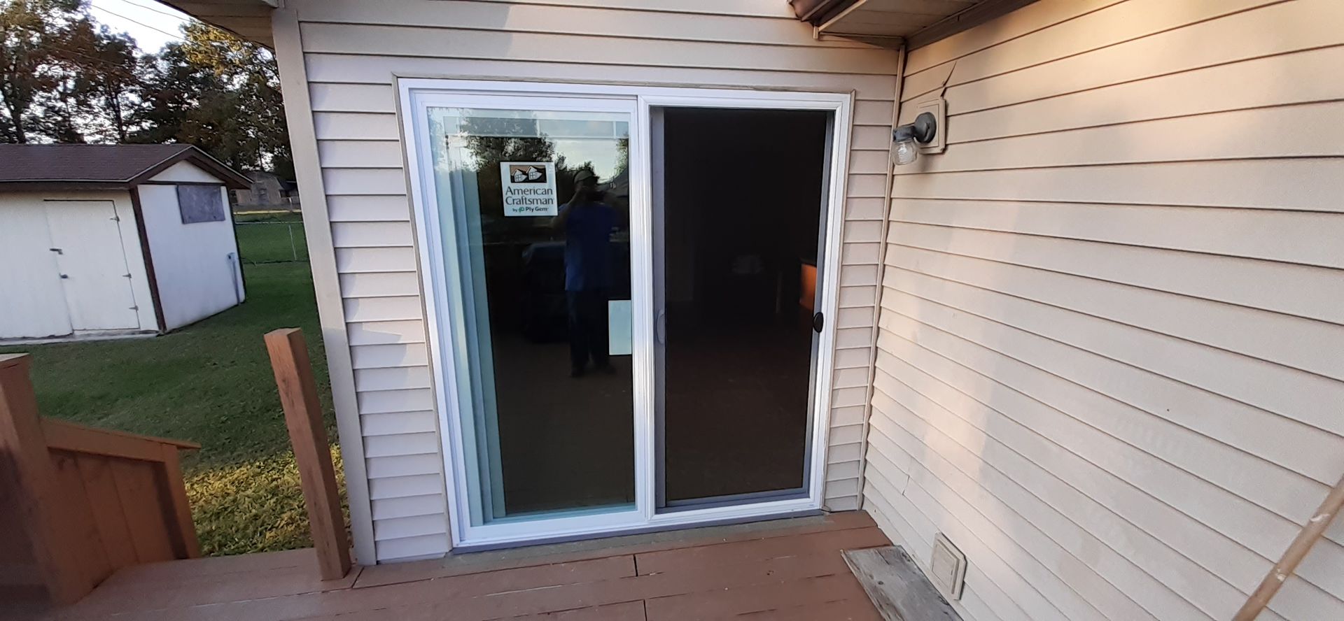 A white sliding glass door installed in the tan horizontal siding of a house, opening onto an outdoor wooden deck.