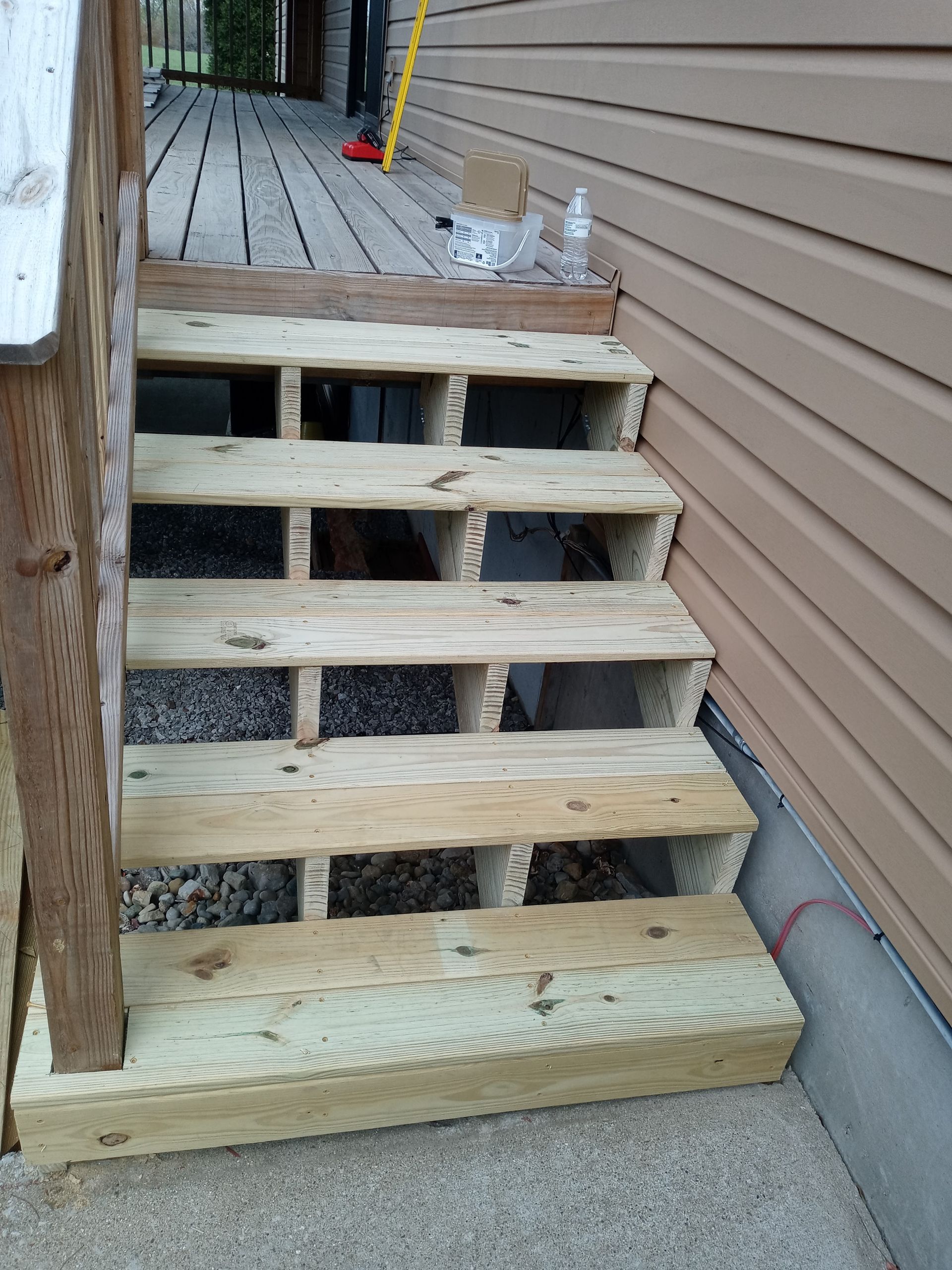 Unfinished wooden steps leading to a deck, showing visible stringers and open risers against a beige house exterior.