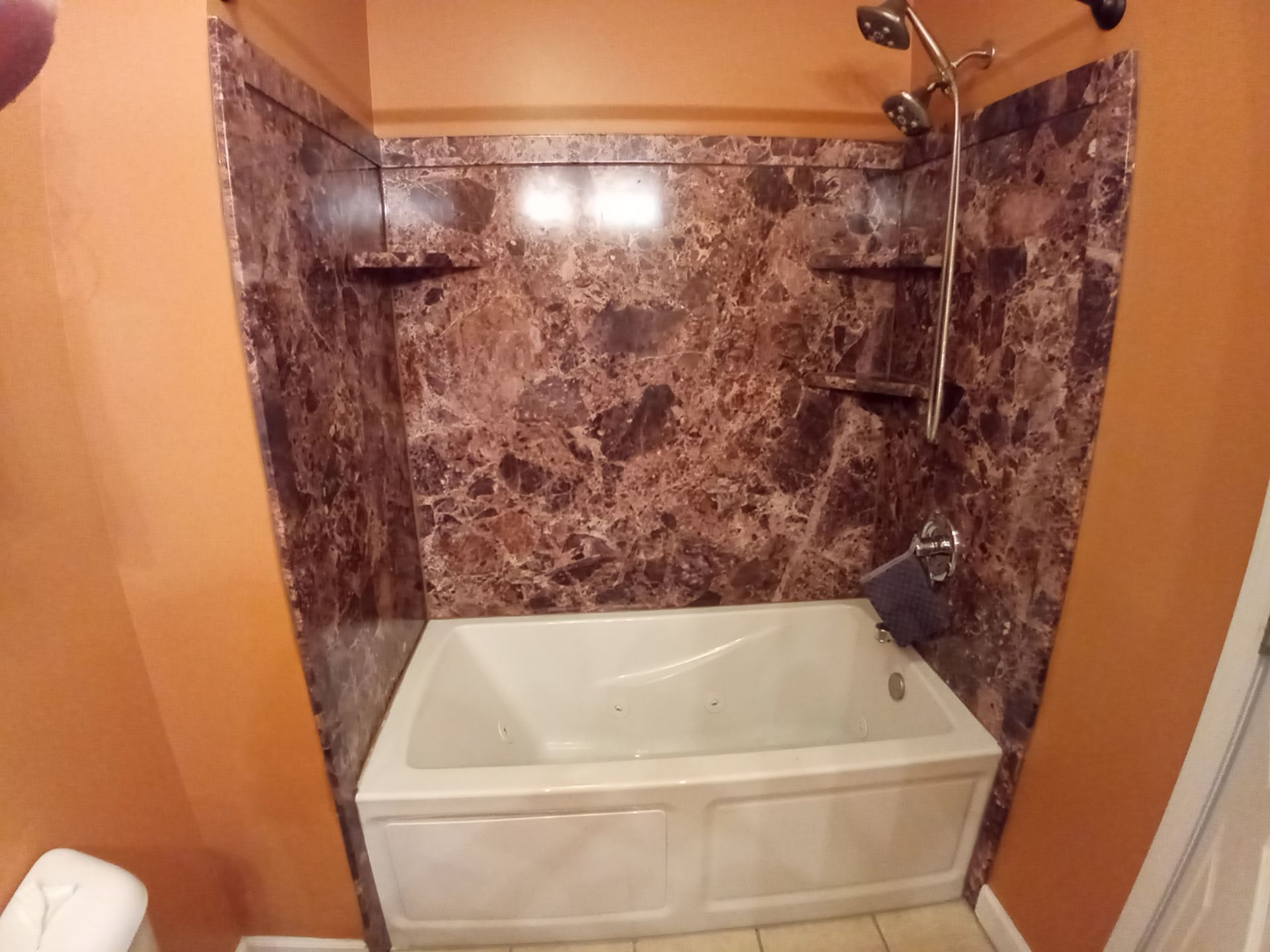 A white bathtub featuring dark brown marble-patterned wall panels with built-in corner shelves and a chrome shower head.