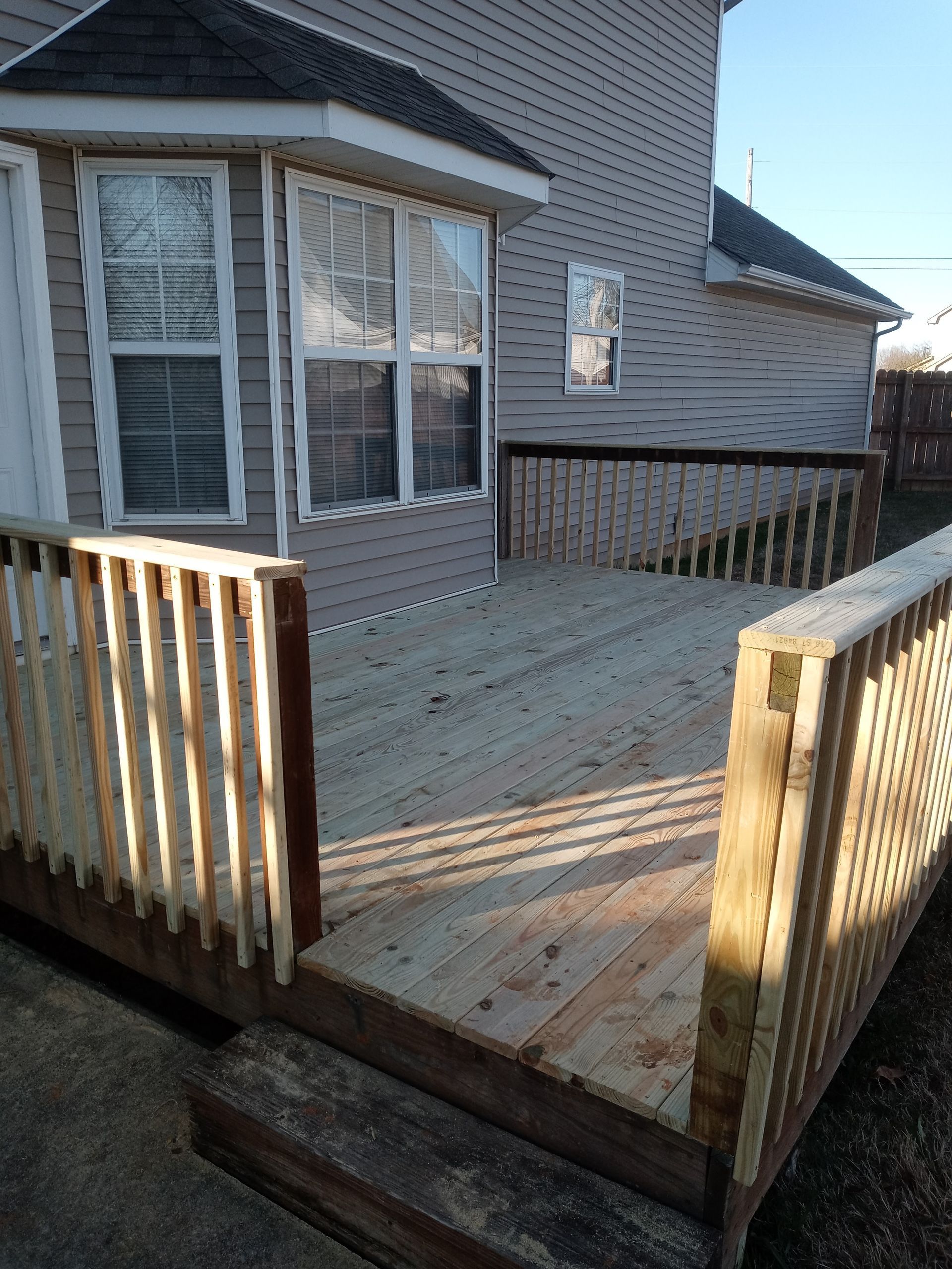 A newly built, light-wood deck with railing attached to the back of a house with tan vinyl siding.