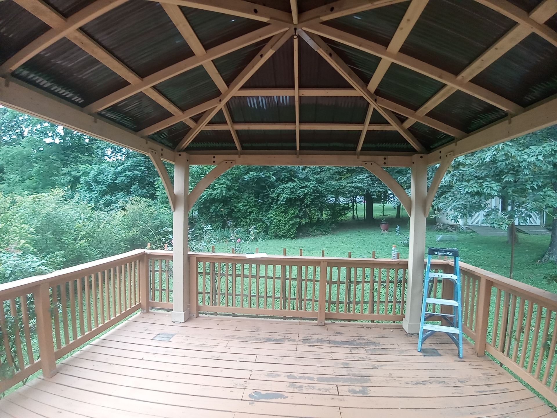 A wooden gazebo structure on an outdoor deck overlooking a grassy, tree-filled yard with a step ladder nearby.