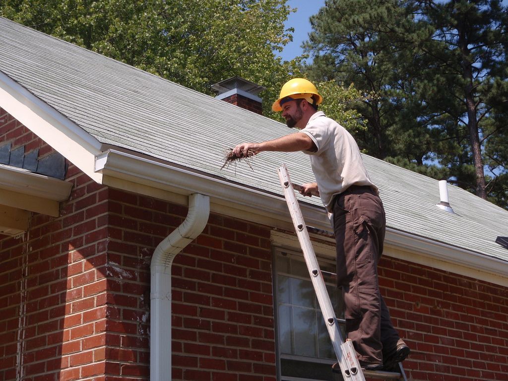 A person in a yellow hard hat standing on a ladder, reaching up to inspect the edge of a shingled roof on a brick house.