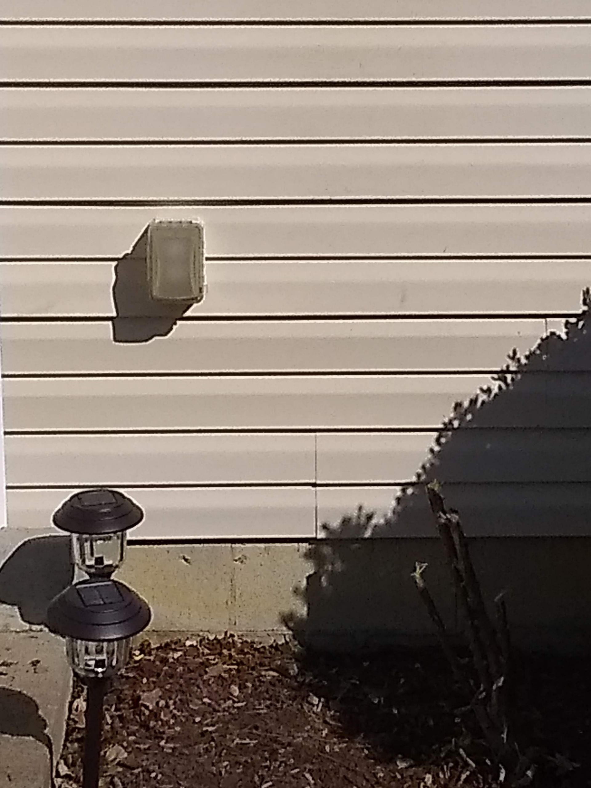 An outdoor view showing light beige horizontal vinyl siding, a square exterior wall vent, and two small garden lights.