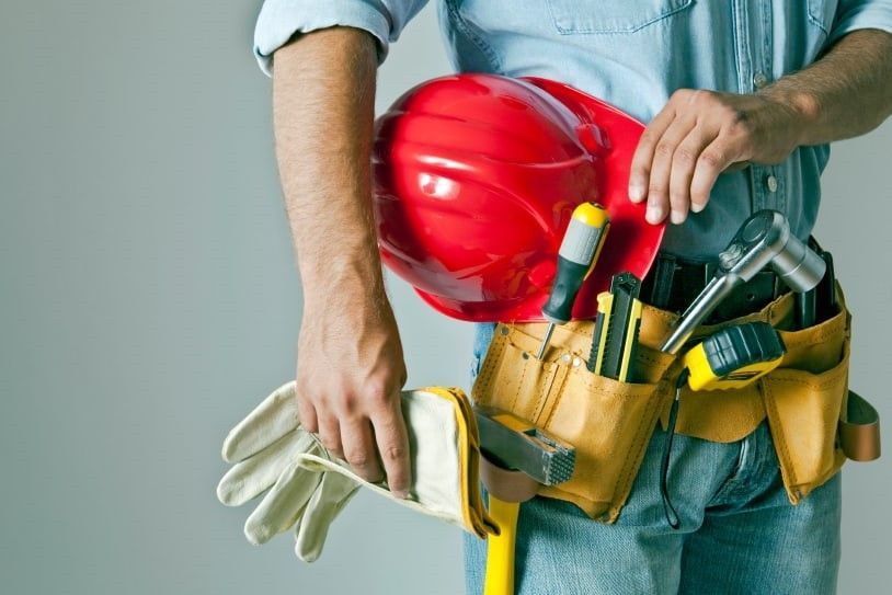 A worker wearing a tool belt with tools, holding a red hard hat and work gloves against a grey background.