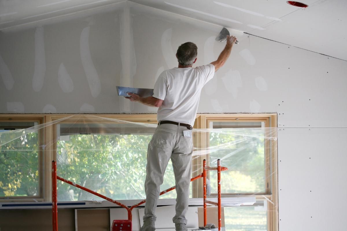 A worker uses a trowel to apply joint compound to the drywall seam of a sloped ceiling in a room under construction.