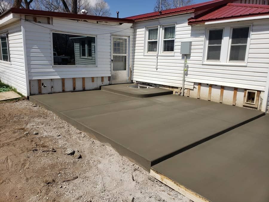 A newly poured concrete patio adjacent to a white house with a red metal roof and unfinished exterior wall trim.