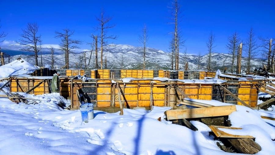 Wooden foundation forms for a building under construction, surrounded by snow, bare trees, and distant snowy mountains.