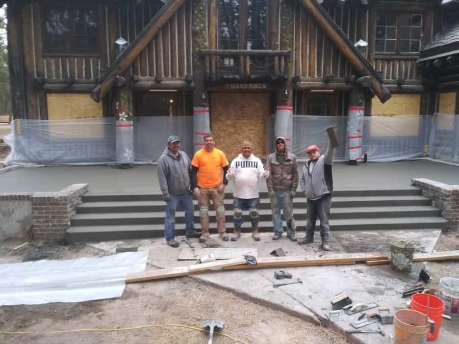 Five construction workers stand in front of a log cabin building on a newly finished concrete porch and stairs.