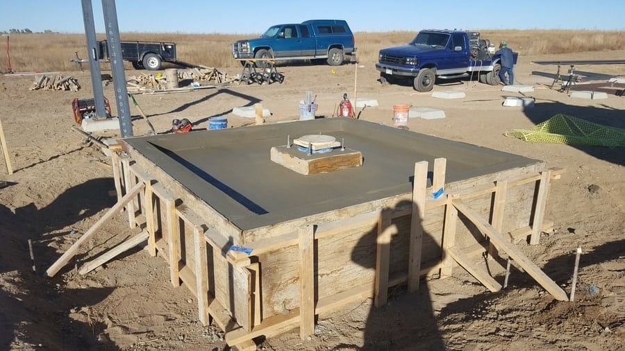 Freshly poured concrete foundation in wooden forms with a central anchor bolt assembly, set in an open, arid field.