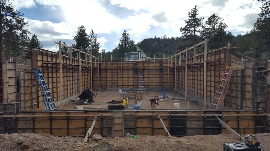 Wooden concrete forms arranged for a building foundation in a construction site surrounded by trees and a cloudy sky.