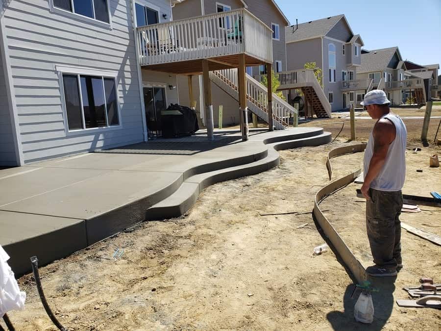 A worker stands on a construction site next to a newly poured, curved concrete patio with built-in steps.