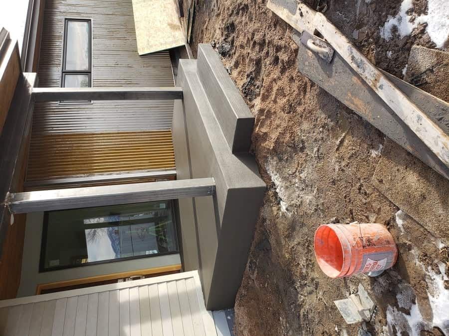 Freshly poured concrete steps at the entrance of a house under construction, with a nearby orange bucket and wooden plank.