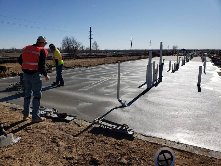 Two construction workers in safety vests finishing a newly poured concrete slab with pipes protruding from the surface.
